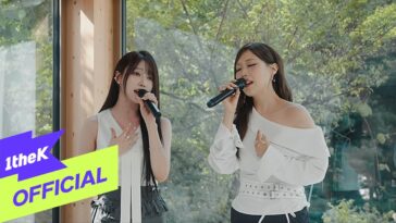 Duas cantoras se apresentam ao vivo, segurando microfones, em um ambiente iluminado por janelas com vegetação ao fundo.