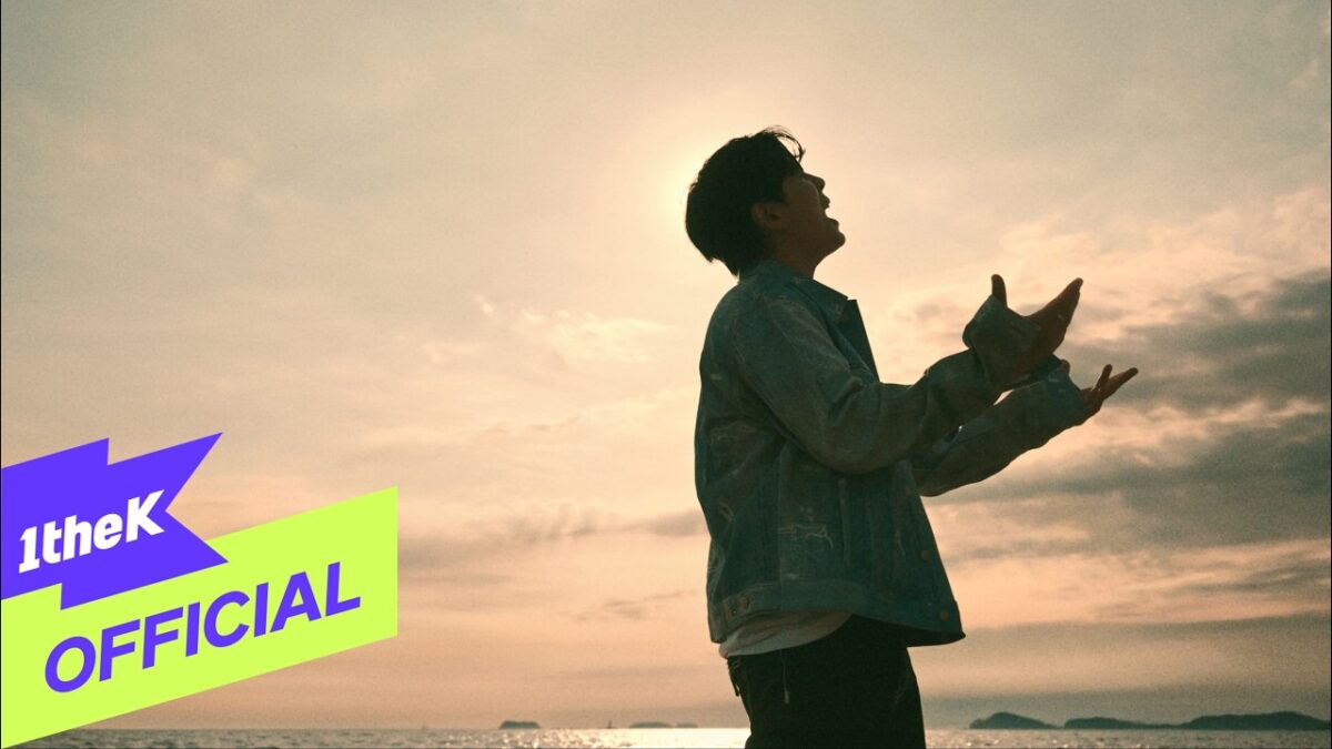 Homem em silhueta canta ao pôr do sol, com o mar ao fundo e nuvens no céu. Logo "1theK" aparece no canto inferior.