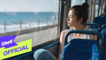 Jovem contemplativa olhando pela janela de um ônibus, com o mar ao fundo em um dia ensolarado.