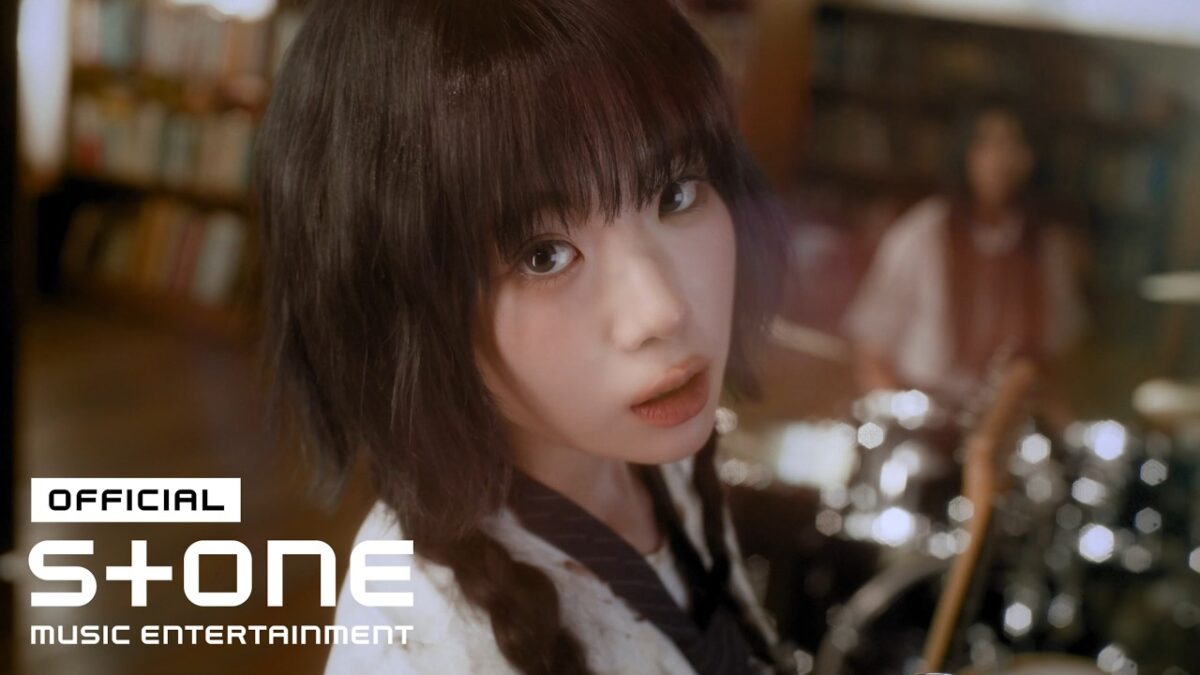 HAENA, com cabelo escuro e tranças, olha intensamente para a câmera em um ambiente musical com instrumentos ao fundo.