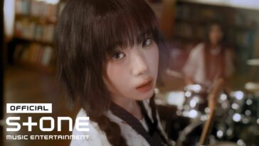 HAENA, com cabelo escuro e tranças, olha intensamente para a câmera em um ambiente musical com instrumentos ao fundo.