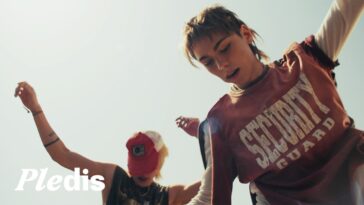 Duas jovens dançam em um ambiente ensolarado, uma usando camiseta "Security Guard" e a outra com boné vermelho.