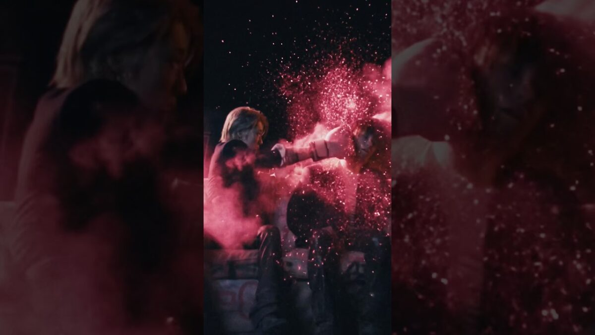 Artista com cabelo loiro agitando os punhos, cercado por uma explosão de pó colorido em cenário dramático.