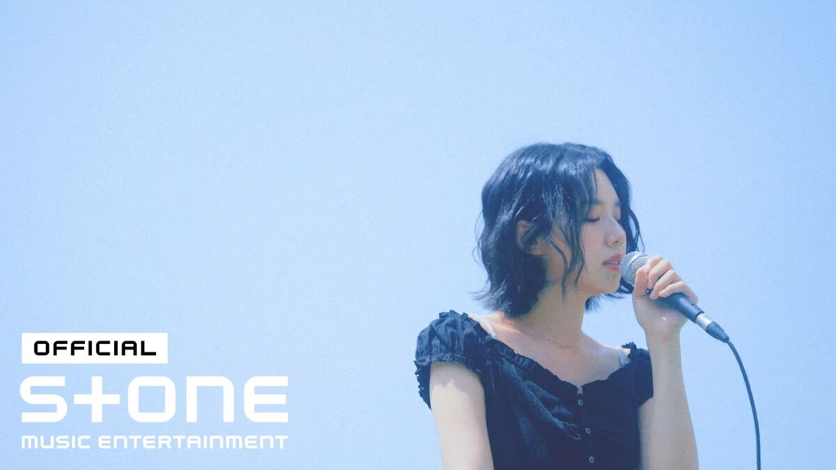 Cantora Choi Ingyeong se apresenta ao vivo, segurando um microfone sob um céu azul claro.