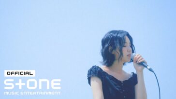 Cantora Choi Ingyeong se apresenta ao vivo, segurando um microfone sob um céu azul claro.