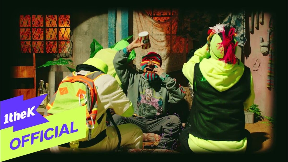 Três artistas em trajes vibrantes interagem em um ambiente colorido e decorado, com um foco em diversão e criatividade.