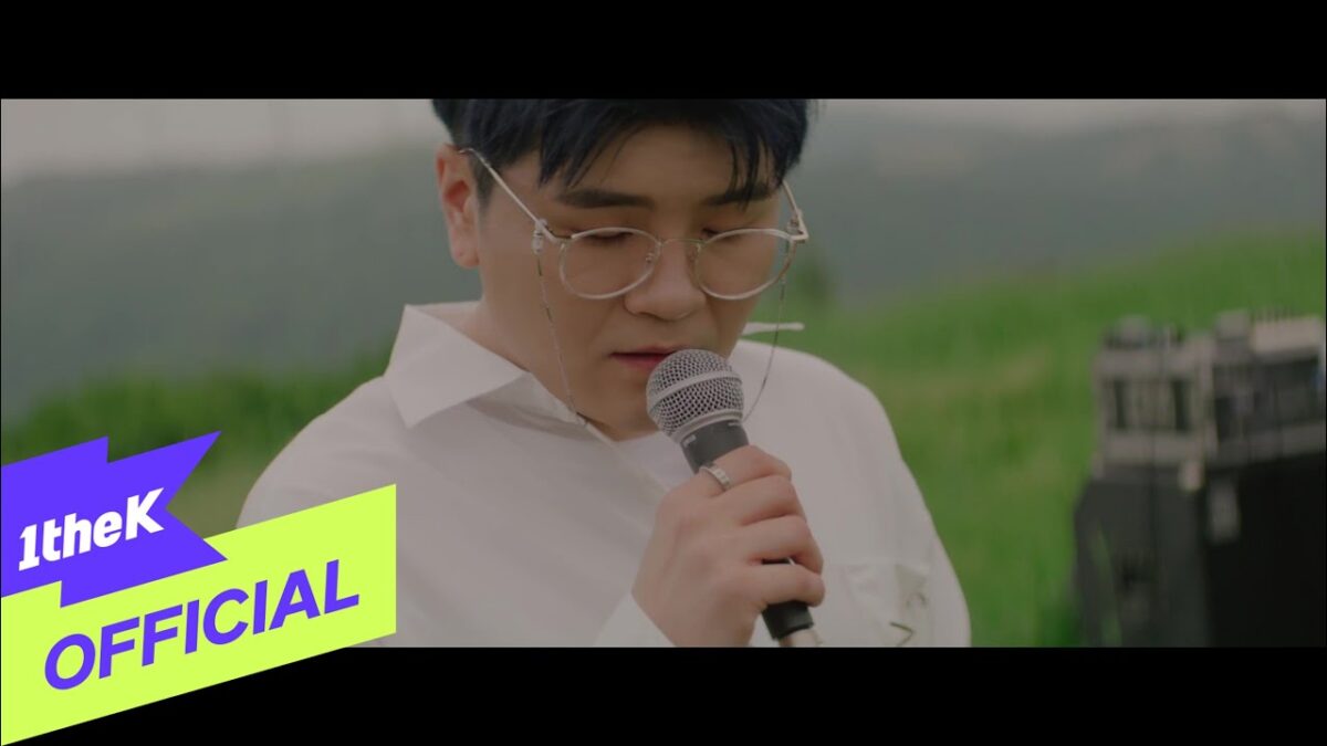Shin Yong Jae canta em um campo verde, segurando um microfone e usando óculos e uma camisa branca.