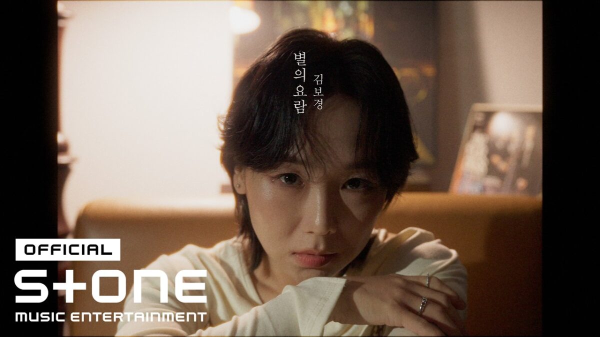 Kim Bo Kyung, com expressão contemplativa, sentada em um sofá, em ambiente com luz suave e elementos decorativos ao fundo.