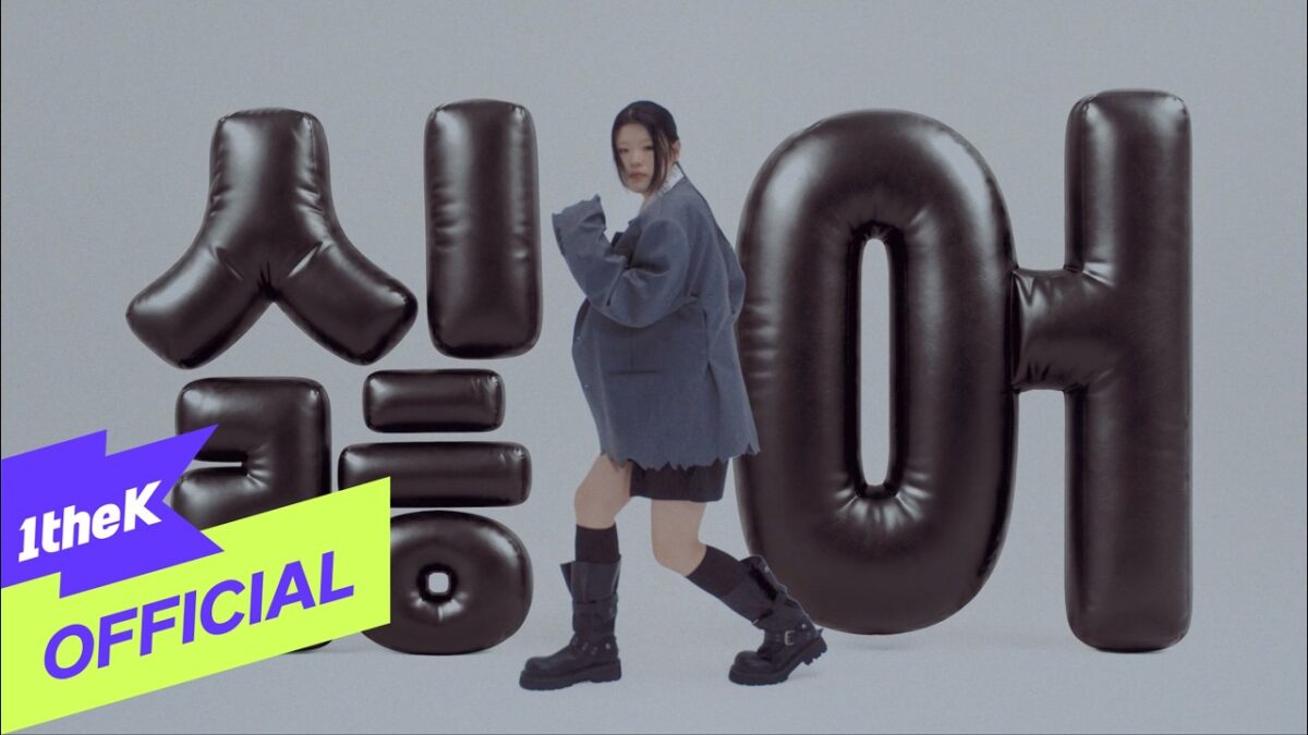 Lee Ye Ji dança em frente a letras infláveis pretas com fundo cinza, vestindo um blazer oversized e botas.