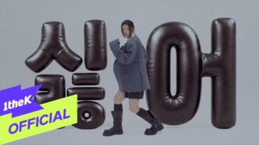 Lee Ye Ji dança em frente a letras infláveis pretas com fundo cinza, vestindo um blazer oversized e botas.