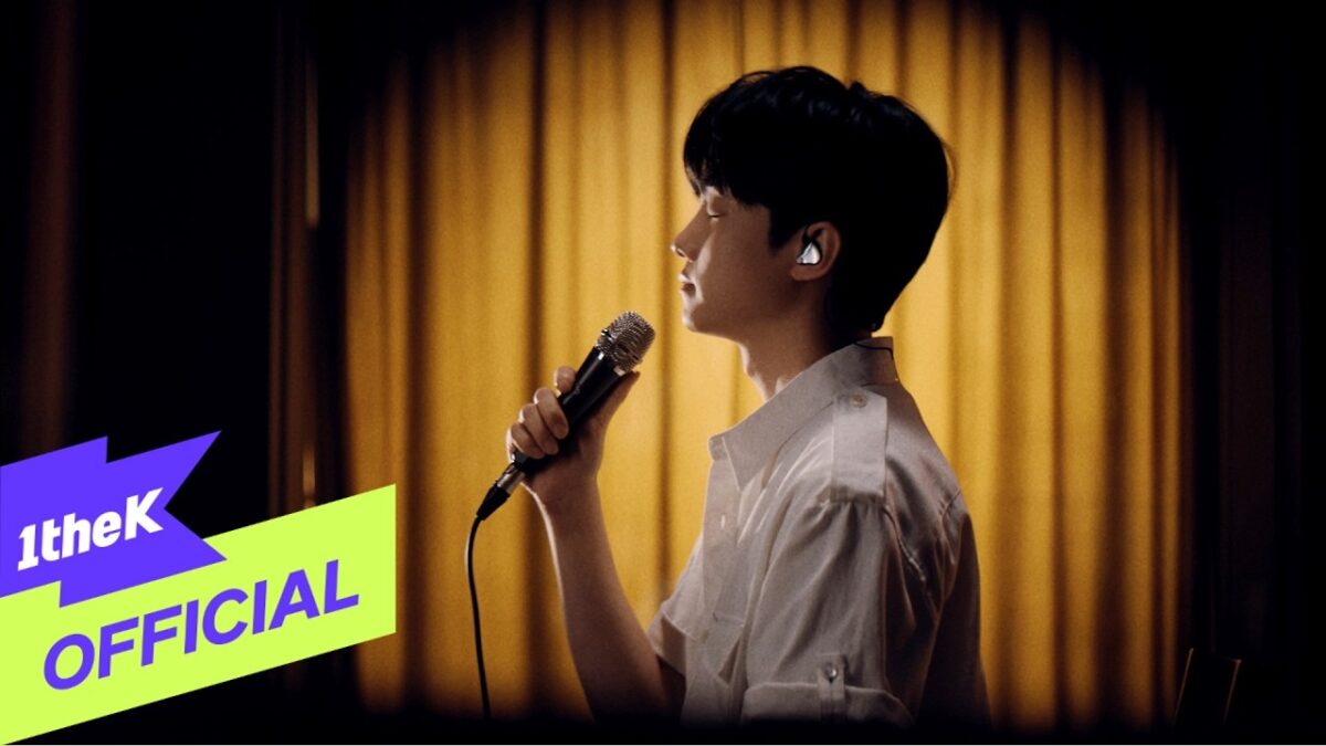 So Soo Bin canta com um microfone em mãos, iluminado por um fundo dourado suave.
