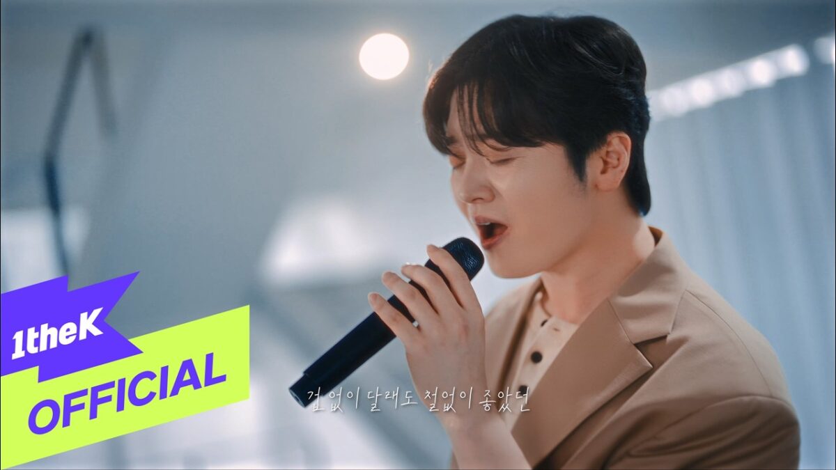 Hwang Karam canta ao vivo, segurando um microfone e expressando emoção em um cenário iluminado.