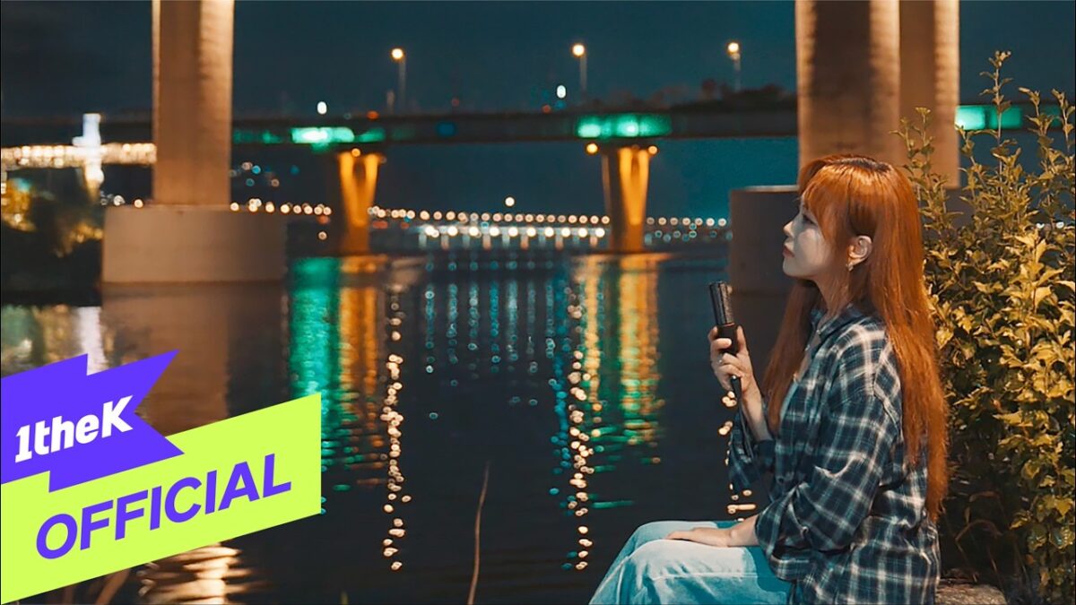 Cantora Lee Ye Joon sentada à beira do rio, segurando um microfone, sob luzes noturnas refletidas na água.