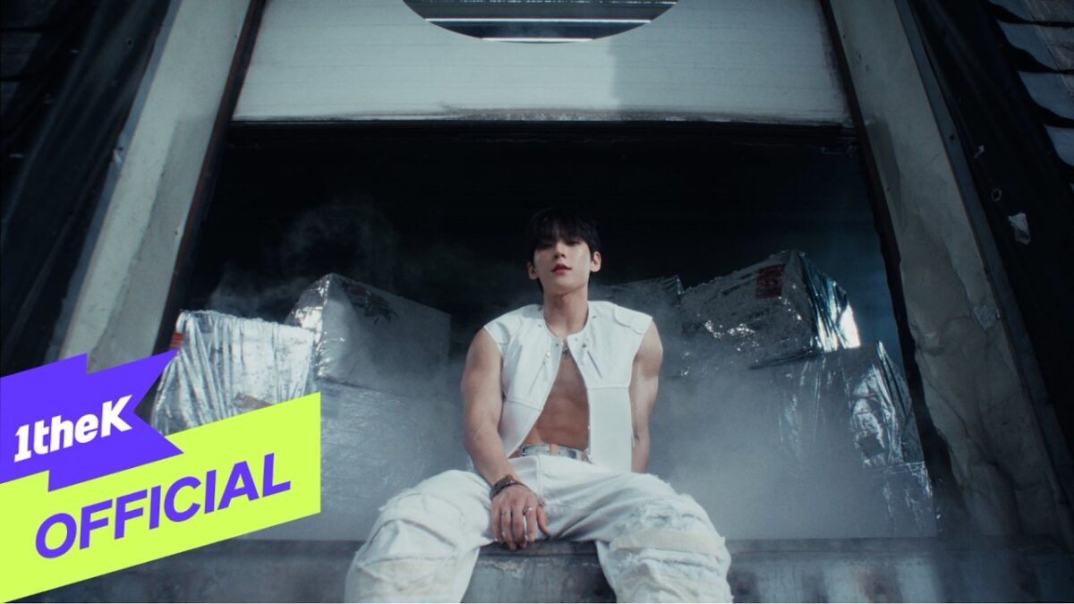 Lee Minhyuk (HUTA) sentado em um cenário com caixas metálicas e fumaça, vestindo uma jaqueta branca sem mangas.
