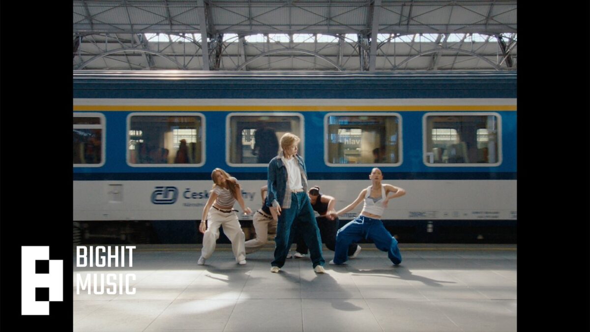 Quatro dançarinos se apresentam em uma estação de trem, com um trem azul ao fundo.