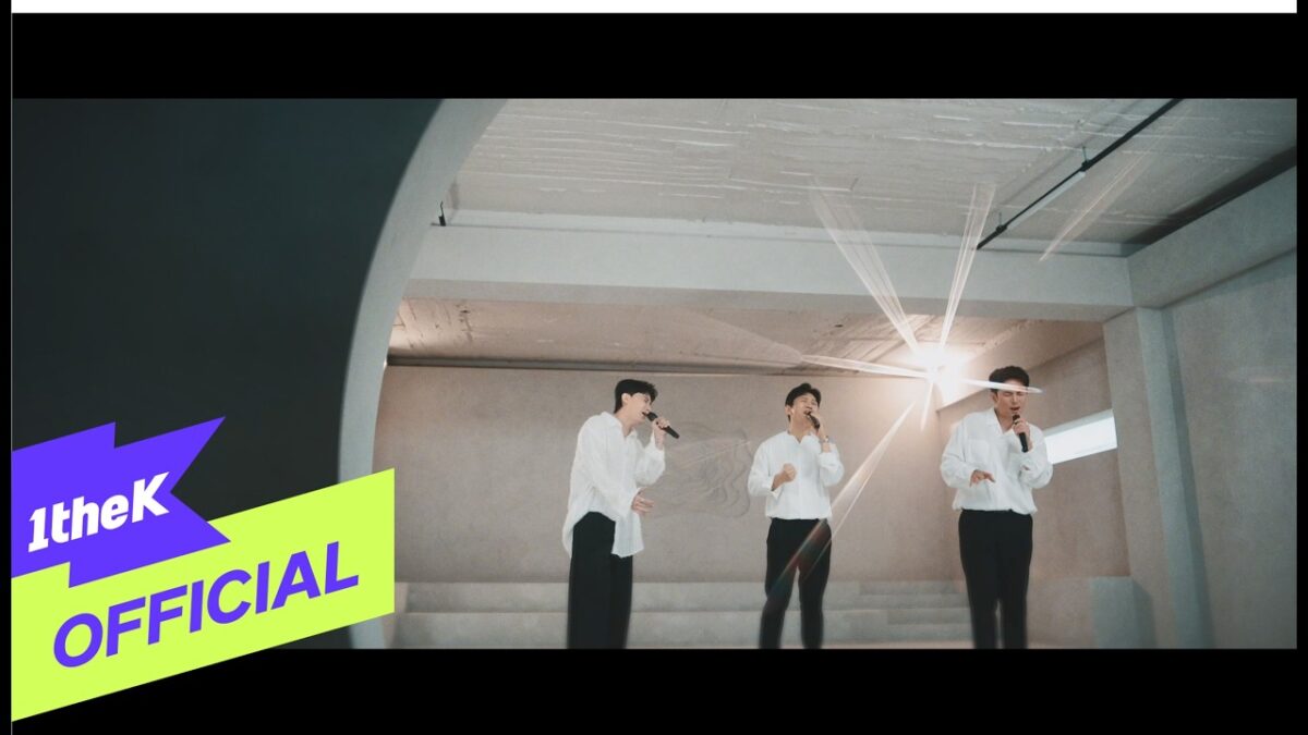 Três cantores em camisas brancas se apresentam em um espaço minimalista, com microfones e iluminação suave.