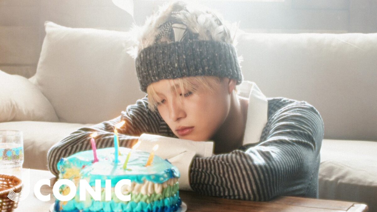 Um jovem com um gorro observa pensativo um bolo de aniversário com velas acesas em um ambiente iluminado.