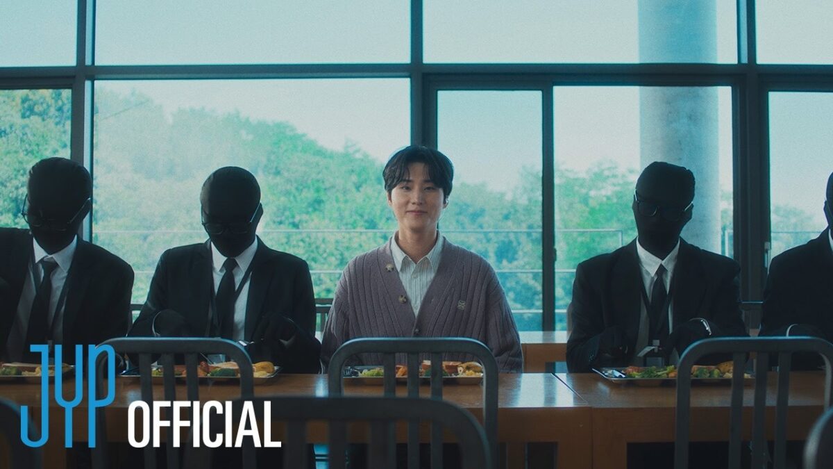 Young K sorri enquanto está sentado em uma mesa com figuras vestidas de preto ao seu redor.