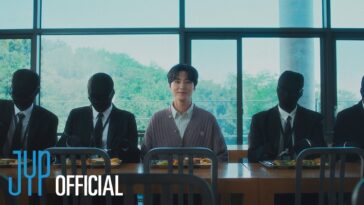 Young K sorri enquanto está sentado em uma mesa com figuras vestidas de preto ao seu redor.