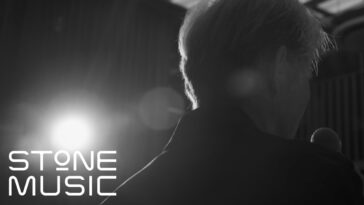 Choi Baek Ho de costas, segurando um microfone sob uma luz suave em ambiente musical.