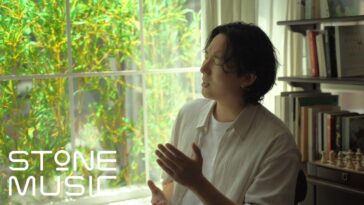 Jae Yeon canta em um ambiente iluminado, com plantas ao fundo e prateleiras de livros ao lado.